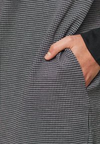 Musta-valkoinen houndstooth-kangas, jossa on texturoitu pinta. Käsi pitää materiaalia, esitellen sen monimutkaisia kuvioita ja laskoksia.