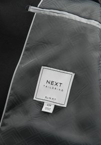 Schwarze maßgeschneiderte Jacke im Inneren mit einem grauen strukturierten Futter. Mit einem weißen Etikett, auf dem "NEXT TAILORING SLIM FIT 40R" sowie Pflegehinweise zu lesen sind.