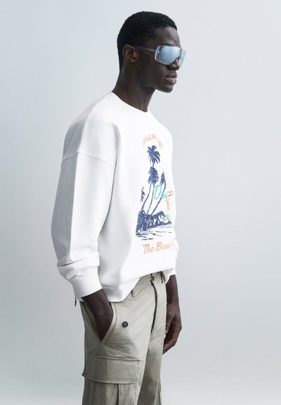 Mann trägt übergroßen weißen Pullover mit Palmenmotiv und beigefarbenen Cargohosen, Hände in den Taschen, große reflektierende Sonnenbrille, Seitenansicht.