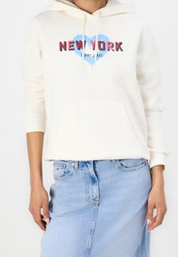 Persona que lleva una sudadera color blanco roto con el gráfico "NEW YORK" y un corazón azul, combinada con una falda de mezclilla azul claro.