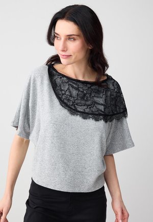 Frau trägt ein lockeres graues Oberteil mit schwarzem Spitzenbesatz im oberen Brustbereich, steht vor einem schlichten hellen Hintergrund.