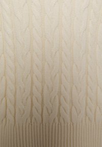 Pull en maille torsadée crème, avec tresses texturées et ourlet côtelé. Le matériau semble doux et chaud, mettant en valeur le design complexe.