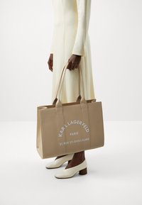 Sac fourre-tout beige avec du texte blanc en relief, doté de poignées solides et d'une forme rectangulaire. Texture lisse, design minimaliste.