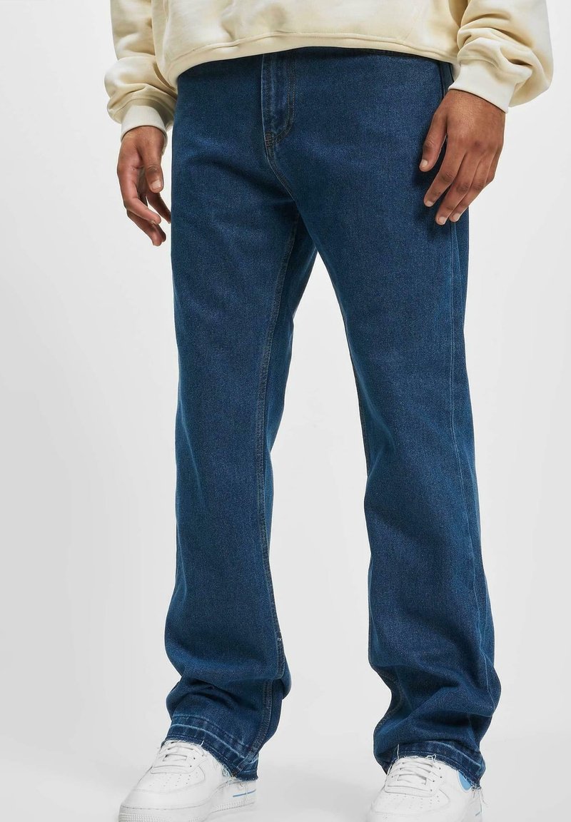 Homme portant un jean bleu foncé ample avec des baskets blanches et un sweat crème visible à la taille et aux poignets.