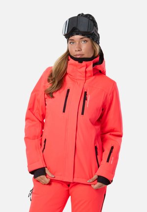 Jeune femme portant une veste de ski corail vif et un pantalon, avec des gants noirs, des lunettes de ski noires sur la tête, debout devant un fond blanc.