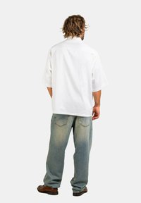 Kurzärmliges weißes Hemd mit einem Rückenpasse, kombiniert mit hellblauen Jeans mit einem verwaschenen Effekt und zwei Gesäßtaschen. Braune Schuhe runden das Outfit ab.