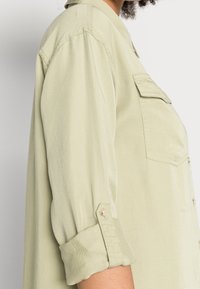 Chemise en boutons vert clair fabriquée en tissu doux. Elle a des manches retroussées, une poche plaquée et un col. Texture lisse avec un léger brillant.