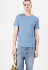 Ανοιχτό μπλε κοντομάνικο μπλουζάκι από απαλό βαμβακερό ύφασμα, συνδυασμένο με ξεθωριασμένα μπλε τζιν. Το μοντέλο κρατάει μια καφέ τσάντα ώμου με διπλές ιμάντες.