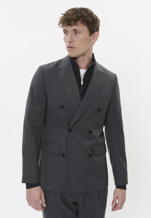 Blazer gris à double boutonnage avec un tissu texturé, des boutons noirs et des revers marqués ; superposé à une veste noire zippée avec une chemise blanche en dessous.