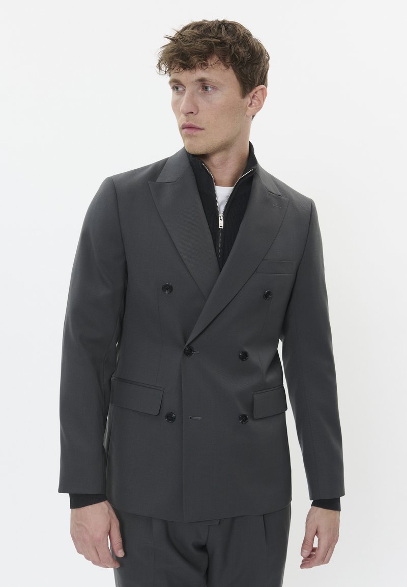 Halli kahepoolse nööbiga bleiser, millel on tekstuuriga kangas, mustad nööbid ja teravad kraed; seljas musta lukuga jakk, mille all on valge särk.