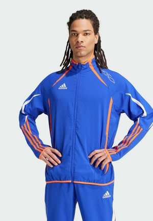 Blauwe sportjack met oranje accenten en witte strepen, rits aan de voorkant en logo op de linkerborst. Lichtgewicht polyester materiaal.