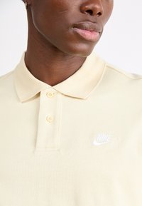 Šviesiai geltona polo marškinėliai, pagaminti iš minkštos medžiagos, su klasikiniu apykakle, dviejų mygtukų užsegimu ir balta siuvinėta Nike ženklu.