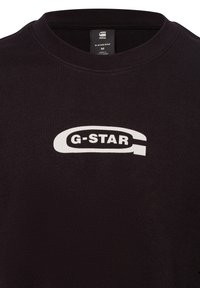 Camiseta de algodón negra con cuello redondo, textura suave y un gran logo blanco "G-STAR" en el frente. Etiqueta de tamaño M visible.