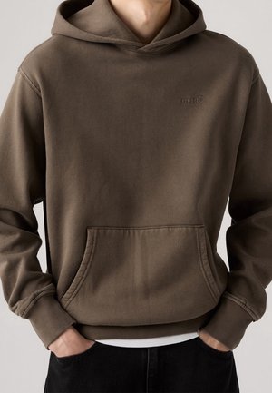 Mężczyzna w brązowej bluzie z kapturem Levi's z przednią kieszenią kangurzą, ręce w kieszeniach i widoczną białą krawędzią podkoszulka.