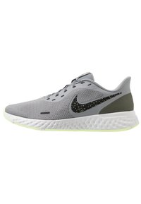 Chaussure de running Nike gris clair avec un swoosh noir à pois, semelle texturée blanche et accent de talon vert olive, vue de côté sur fond blanc.
