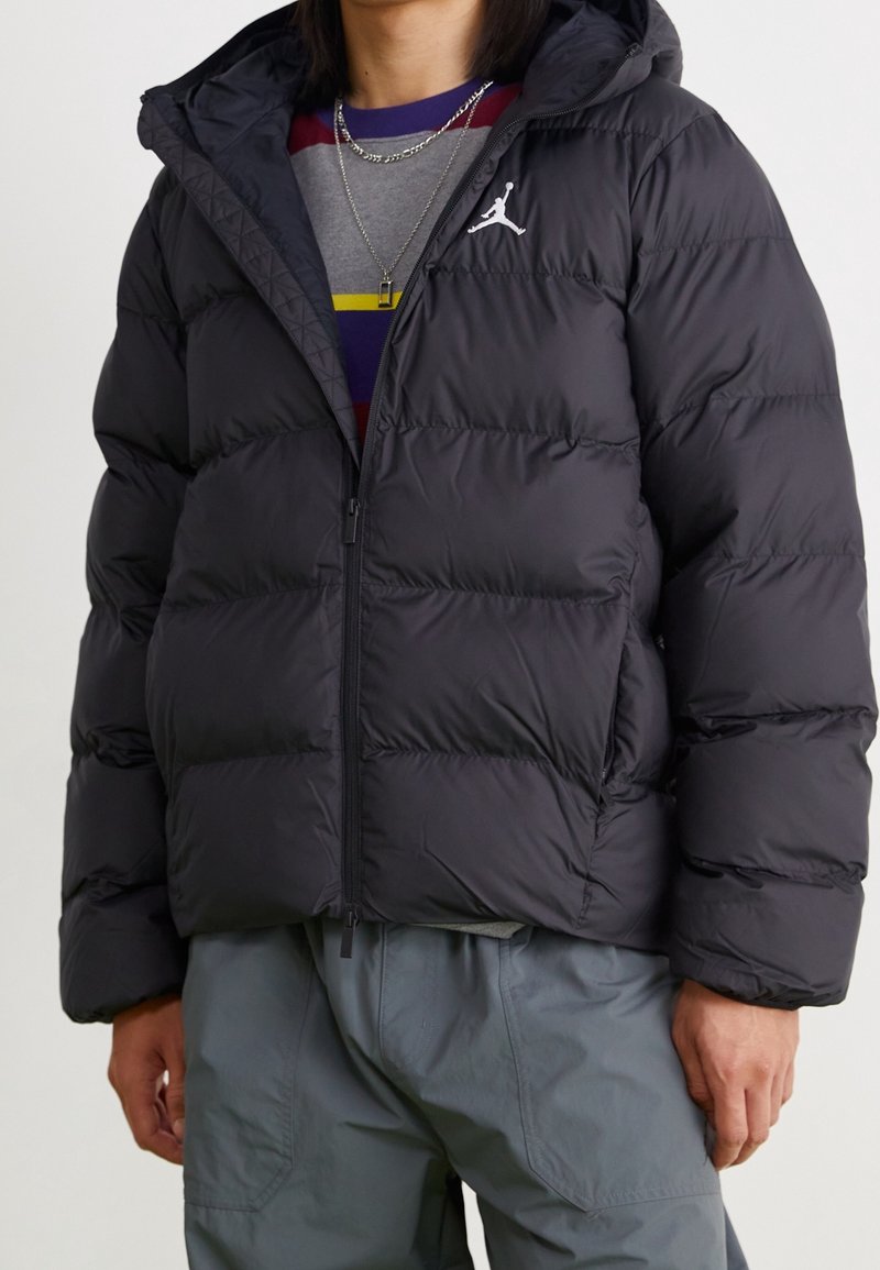 Schwarze Puffjacke mit Kapuze, ausgestattet mit einem gesteppten Design, vorderem Reißverschluss und dem Jordan-Logo auf der linken Brust. Getragen über einem grauen Shirt.