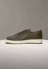 Brązowy, niskoprofilowy sneaker ze skóry, z białą gumową podeszwą, sznurowanym zapięciem i gładkim wykończeniem, prezentowany na jasnoszarej powierzchni.