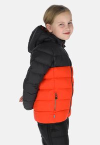 Kind draagt een zwart en fel oranje gewatteerd jack met capuchon, staat in profiel met handen achter de rug, glimlacht lichtjes.