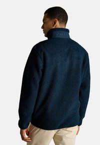 Mies, który ma na sobie granatową polarową kurtkę z wysokim kołnierzem i wytłoczonym logo "JOULES" na plecach, w połączeniu z beżowymi spodniami, odwrócony plecami.