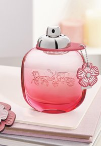 Pink glass perfume bottle with rounded edges, silver cap, embossed horse-drawn carriage design, and a pink flower-shaped tag attached.