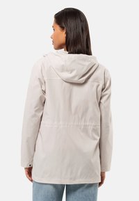 Helle beige Kapuzenjacke mit glatter Textur, sichtbaren Nähten, verstellbarem Kordelzug und lockerer Passform. Dazu gehören blaue Jeans.