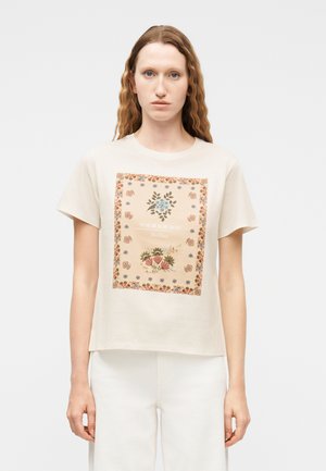 Frau mit langen, welligen Haaren, die ein beiges T-Shirt mit floralem Quadrat-Print und dem Schriftzug "Weekend" trägt, kombiniert mit einer weißen Hose.