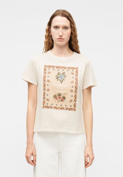 Femme aux longs cheveux ondulés portant un t-shirt beige avec un imprimé carré floral et le texte "Weekend", associé à un pantalon blanc.