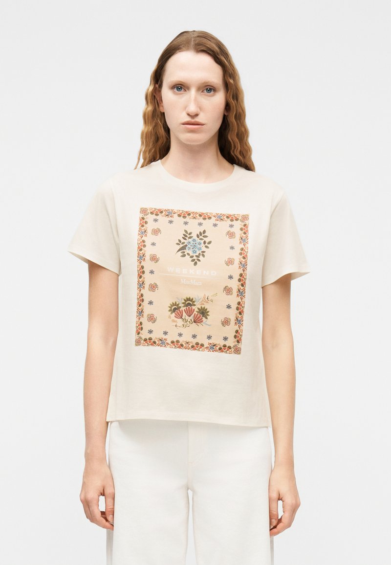 Γυναίκα με μακριά κυματιστά μαλλιά που φοράει μπεζ μπλουζάκι με τετράγωνο λουλουδάτο σχέδιο και το κείμενο "Weekend", σε συνδυασμό με λευκό παντελόνι.