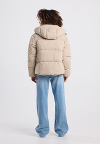 Giacca puffa beige con cappuccio, design trapuntato e vestibilità rilassata, abbinata a jeans a gamba larga blu chiaro. Texture liscia, dettagli minimi.