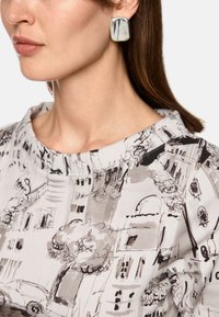 Gray blouse with black line drawings of buildings; model wears white and gray marbled clip-on earrings. Smooth texture, rounded shapes.