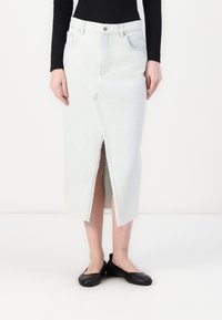 Jupe en denim bleu clair avec fente avant, longueur mi-mollet, bord effiloché et design traditionnel à cinq poches, associée à des chaussures plates noires.