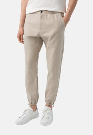 Beige, tapered bukser med elastiske manchetter og knap foran, båret med hvide sneakers og en hvid skjorte, hænder i lommerne.