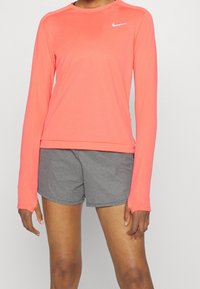 Maglietta a maniche lunghe Nike di un vivace corallo, realizzata in tessuto morbido, abbinata a pantaloni sportivi grigi. Presenta un collo rotondo e dettagli con il logo.