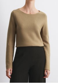 Femme portant un haut en tricot beige à manches longues associé à un pantalon noir taille haute, debout devant un fond blanc uni.