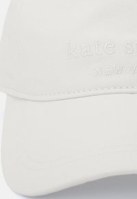 Vit keps i texturerad tyg med upphöjd brodering av "kate spade NEW YORK" framtill, med en rundad skärm och sömdekor.