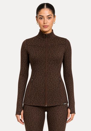 Femme portant une veste zippée marron ajustée à imprimé léopard et un pantalon assorti avec des trous pour les pouces, debout devant un fond clair uni.