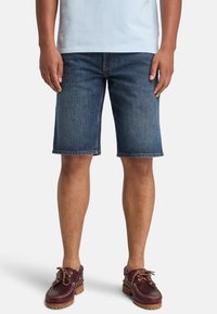 Pantalones cortos de mezclilla en azul oscuro con un corte recto, dobladillo deshilachado y costuras naranjas en contraste, combinados con zapatos de barco de cuero marrón.