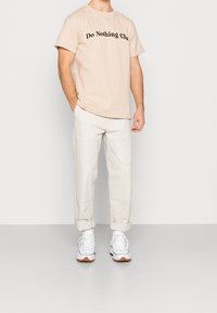 Muž v béžovém tričku "Do Nothing Club", světlých krémových kalhotách s ohrnutými manžetami, bílých vysokých teniskách, stojící s jednou rukou v kapse.