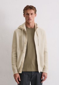 Cremefarbener gestrickter Cardigan mit Kapuze, der einen durchgehenden Reißverschluss und Knöpfe hat. Über einem hellolivfarbenen T-Shirt und dunklen Hosen getragen.