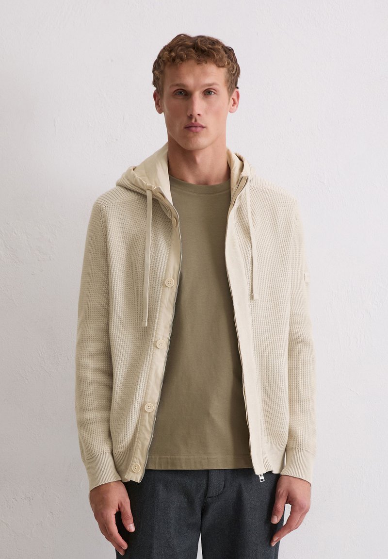 Cremefarbener gestrickter Cardigan mit Kapuze, der einen durchgehenden Reißverschluss und Knöpfe hat. Über einem hellolivfarbenen T-Shirt und dunklen Hosen getragen.