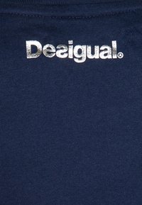Srebrny, refleksyjny nadruk logo "Desigual" na ciemnoniebieskiej, teksturowanej tkaninie, prawdopodobnie część odzieży.