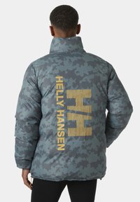 Přifukovací bunda v šedém camouflage vzoru s vysokým límcem. Má velké žluté logo "Helly Hansen" na zádech a hladký povrch.