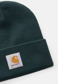 Mørkegrønn ribbet strikket lue med brettet oppkant som har en hvit etikett med et oransje Carhartt-logo og rødt tekst.