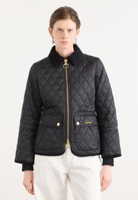 Zwarte gewatteerde jas met een gouden ritssluiting, drukknoopzakken en een ribstoffen kraag. Heeft een getailleerd ontwerp en contrasterende manchetten. Minimale branding zichtbaar.