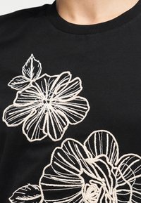 Gros plan sur un tissu noir avec des motifs de fleurs et de feuilles brodés en blanc sur un vêtement à col rond.