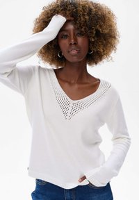 Pull en maille blanche avec un décolleté en V, présentant une texture à motifs près du col, des manches longues et une coupe décontractée. Assorti à un jean bleu.