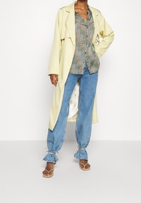 Light yellow trench coat over a patterned button-up shirt, paired with loose blue jeans featuring ruffled cuffs, and tan sandals.