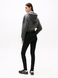 Kvinna i grå kortärmad hoodie, svarta tajta jeans och svarta sneakers står med ryggen vänd och huvudet lutat åt sidan mot en vit bakgrund.