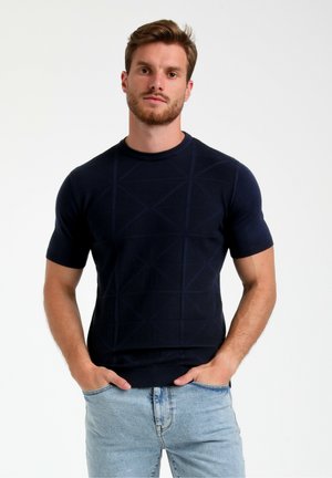 Suéter de punto de manga corta azul marino con un patrón geométrico y cuello acanalado. Llevado con jeans de color azul claro, con las manos en los bolsillos.
