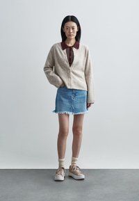 Beige strikket cardigan med ribbet tekstur, kombineret med en vinrød skjorte med krave, denim mininederdel med frynset kant og lysebrune sneakers.